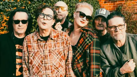 The band pose outside with a wall and greenery behind them. Three have sunglasses on and two are wearing checked shirts with another in a black jacket and a fourth member in a green jacket. Two others are obscured in the photo.