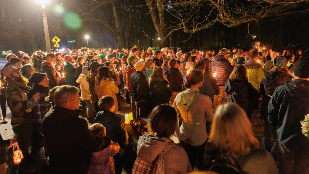 Hundreds of people are shown facing away from the camera holding candles in a vigil. They are wearing a variety of winter gear.