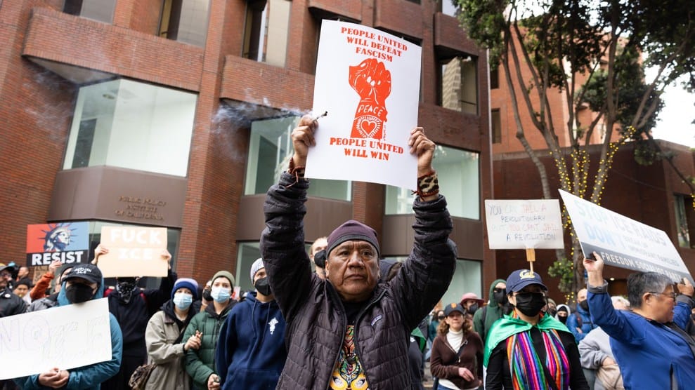 A man stands among a crowd, holding a sign over his head that that reads, "People united will defeat fascism, people united will win."