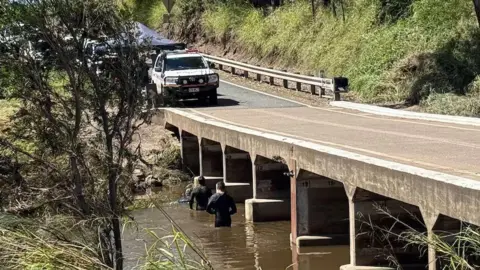 Two men in wet suits wade in a brown river running underneath a concrete road bridge with a rescue vehicle and tent on the road