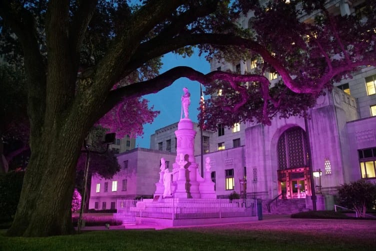 Pink light illuminates a statue, which includes a soldier and the busts of several men, in front of a large government building.