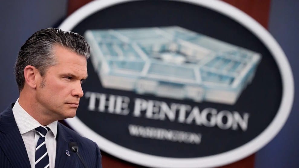 A white man with slick back hair, Pete Hegeseth, stands in front of the Pentagon logo.